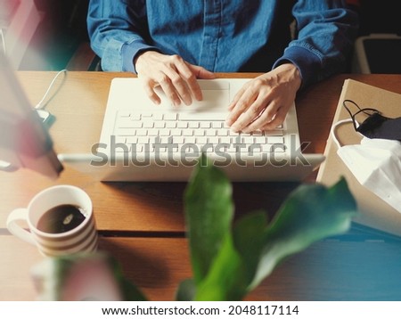 Work from home during the COVID-19 pandemic theme male hands using laptop on wooden table with double mask over paper file folder and blurred green plant in foreground. (selective focus on hand)