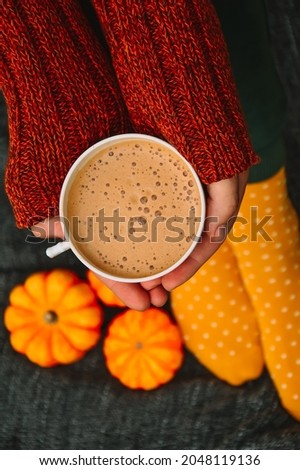 A woman in a brown knitted sweater and yellow socks is holding a cup of cappuccino coffee against a gray blanket. A warm and cozy autumn. Hugge. Peace and comfort. Yellow-orange pumpkins. Autumn mood