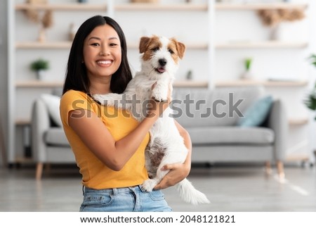 Portrait of cheerful young asian woman in casual outfit carrying her sweet fluffy dog jack russel terrier breed, pet and owner having good time together at home, living room interior, copy space Royalty-Free Stock Photo #2048121821