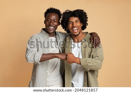 Portrait Of Two Cheerful Black Guys Embracing And Making Fist Bump Gesture, Happy Young African American Male Friends Having Fun Together While Standing Over Beige Studio Background, Copy Space