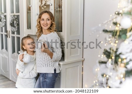 family mother and child daughter in warm white sweaters drinking tea and smiling in decorated living room