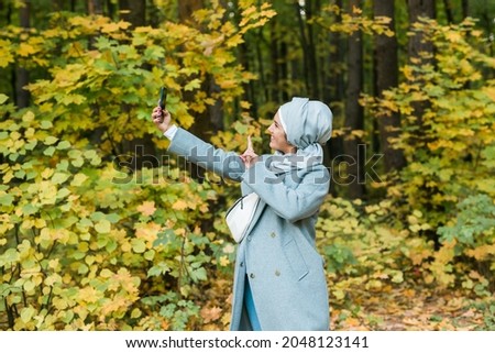 Fashionable young muslim asian girl in hijab taking a selfie on smartphone outdoors in autumn park.