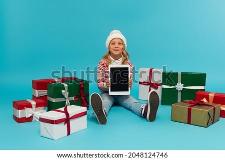 happy kid in jumper and hat sitting with digital tablet near multicolored gift boxes on blue