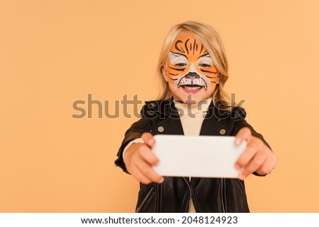 smiling girl in tiger makeup taking selfie on blurred smartphone isolated on beige