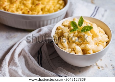 Mac and cheese in white bowl with basil on top and another mac and cheese baked in oven in background placed on a white rustic board Royalty-Free Stock Photo #2048127839