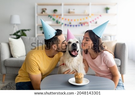Young interracial couple in party hats celebrating their dog's birthday, kissing their pet at home. Millennial owners presenting golden retriever with small b-day cake indoors