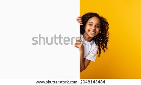 Intresting Offer. Happy Casual Black Girl Peeping Out The Side Of White Advertisement Board For Text Or Design. Smiling Cheerful Teenage Holding Billboard, Looking At Camera, Standing Over Yellow Wall