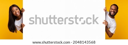 Promotion Billboard Concept. Cheerful African American Guy And Lady Holding White Advertisement Board, Standing Together On Yellow Background, Looking At Camera Peeping Out The Two Sides Of Sign Board Royalty-Free Stock Photo #2048143568