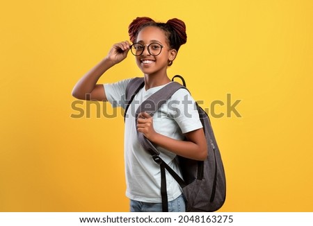 Happy pretty african american school girl wearing glasses, holding backpack on yellow studio background, smart black schooler going to school, enjoying studying. School, education concept Royalty-Free Stock Photo #2048163275