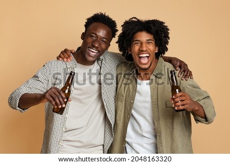 Portrait Of Two Happy Black Friends Embracing And Holding Bottles With Beer, Cheerful African American Guys Having Fun While Posing Together Over Beige Background In Studio, Enjoying Party