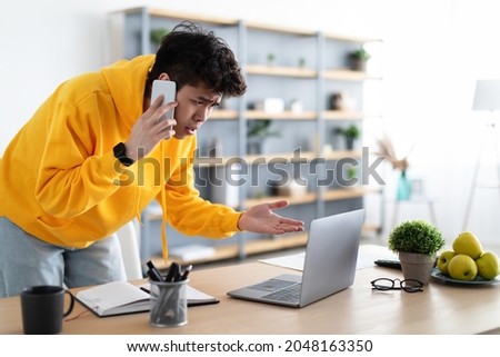 Confused Discontented Asian Man Talking On Phone Using Laptop Having Problem With Computer Or Internet Connection At Home. Displeased Male Customer Calling To Hotline Service And Complaining Royalty-Free Stock Photo #2048163350