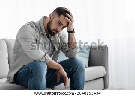 Man sit alone feeling sad worry regret or fear, depressed and desperate in sofa at home. Suffering emotional pain and unhappy, mental health. Young european handsome guy presses hand to forehead Royalty-Free Stock Photo #2048163434