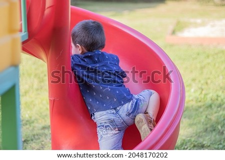 Happy handsome 3 year old boy riding a red slide in an outdoor playground, near a forest. Nature background. The kid is dressed in jeans short pants and blue shirt with white anchors.summertime