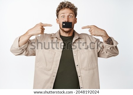 Funny blond guy holds credit card in his mouth and pointing fingers, recommending bank, open account here, standing over white background