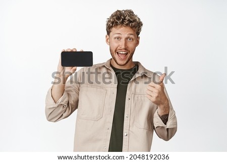 Happy young man shows horizontal phone screen and thumbs up, recommending smartphone app, pleased with awesome application, white background