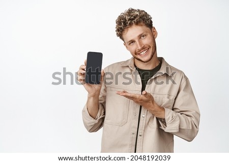 Smiling blond man demonstrating a mobile app, holding smartphone and showing on smartphone screen, standing over white background