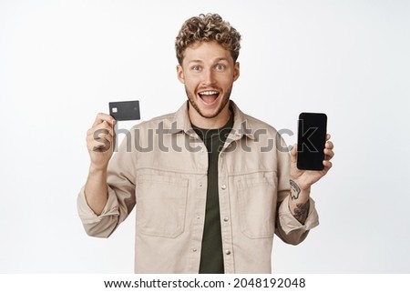 Young happy blond man showing credit card with mobile phone screen, smiling amazed, announcing awesome promotion, standing over white background