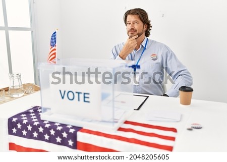 Handsome middle age man sitting at voting stand looking confident at the camera with smile with crossed arms and hand raised on chin. thinking positive. 