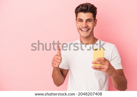 Young caucasian man holding a mobile phone isolated on pink background smiling and raising thumb up