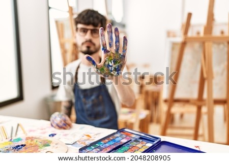 Hispanic man with beard at art studio doing stop sing with palm of the hand. warning expression with negative and serious gesture on the face. 