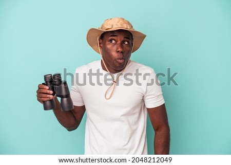 Young African American man holding binoculars isolated on blue background shrugs shoulders and open eyes confused.