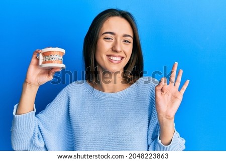 Young hispanic girl holding orthodontic doing ok sign with fingers, smiling friendly gesturing excellent symbol 