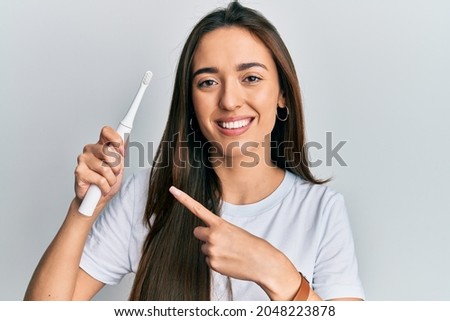 Young hispanic girl holding electric toothbrush smiling happy pointing with hand and finger  Royalty-Free Stock Photo #2048223878