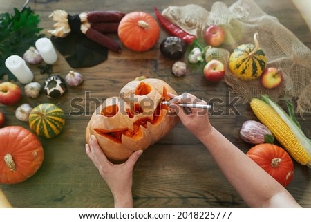 Halloween concept: top view of female hands carving pumpkin. Woman carves a mouth in a pumpkin to make a jack o'lantern on a wooden table with vegetables. High quality image
