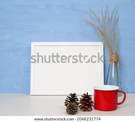 White frame mockup for design or text,red coffee mug,dried palm leaf in glass vase and pine cones  on a wooden table blue wall background