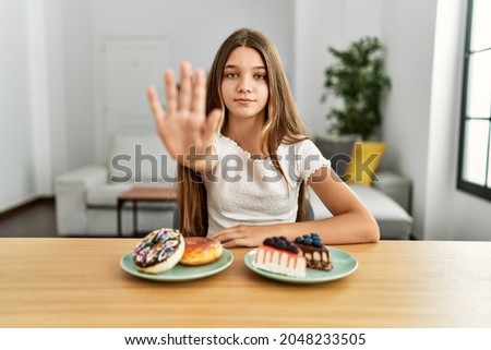 Young brunette teenager eating cakes and sweets with open hand doing stop sign with serious and confident expression, defense gesture 