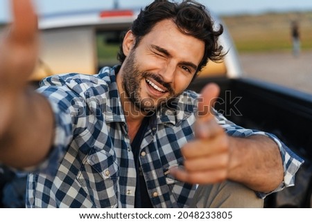 Young white man smiling and taking selfie while sitting in trunk during journey