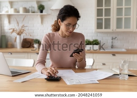 Happy young Caucasian woman sit at desk at home manage household expenses expenditures paying on cellphone online. Millennial female calculate budget finances, make payment on smartphone. Royalty-Free Stock Photo #2048241059