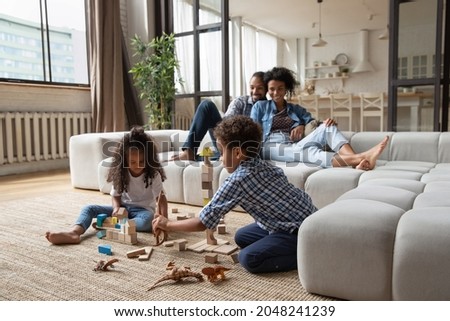 Little African American kids playing with wooden construction toys while parents relaxing on couch, 5s girl and boy sitting on warm floor in living room, having fun together, enjoying weekend Royalty-Free Stock Photo #2048241239