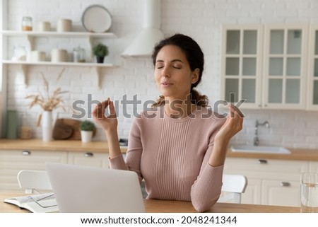 Calm millennial Caucasian female sit at desk at home office work on laptop meditate with mudra hands. Young woman distracted from computer job practice yoga breathe fresh air. Peace concept.