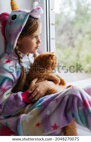 ginger kitten and a little girl in a kigurumi sit on the windowsill and look out the window. autumn, children.