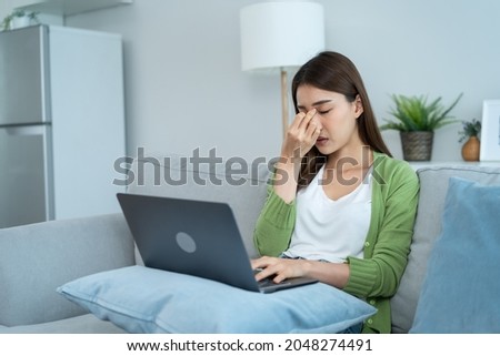 Exhausted Asian business woman hurt eye while using laptop computer. Young girl office worker sit on sofa feel visual fatigue and eye strain tired from overwork and massage dry irritable eye at home. Royalty-Free Stock Photo #2048274491