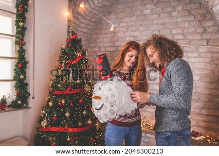 Young couple in love decorating home for Christmas, making a snowman by joining white plastic cups with stapler; people having fun making Christmas decorations