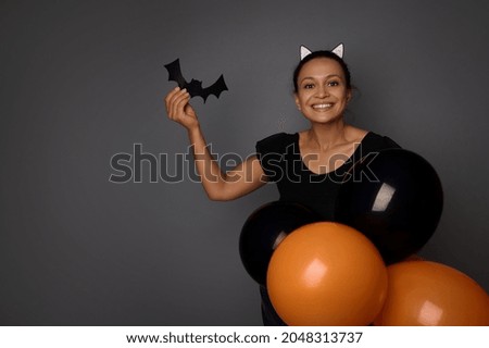Beautiful Hispanic woman dressed in Halloween carnival costume smiles looking at camera, holds handmade bat in her hand , poses with colorful black orange air balls against gray background, copy space