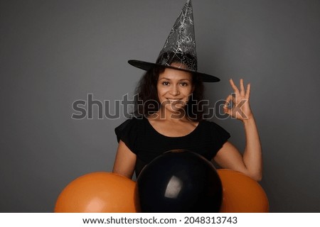 Beautiful smiling woman in wizard hat, dressed in a witch carnival costume for Halloween party, shows OK sign, poses against gray background with black balloons, copy space