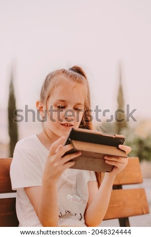 Teen girl wearing casual tee, sitting on wooden bench in park alone, holding gadget tablet pad in hands. Having videocal, playing games. Kids using technologies