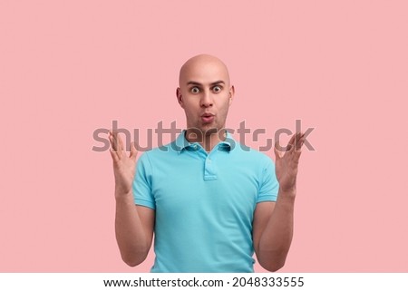 Impressed bald homosexual man with bristle shows big size with both hands, shapes huge object, has eyes wide open, pulls lips, gay friendly, wears blue shirt, stands over pink background Royalty-Free Stock Photo #2048333555
