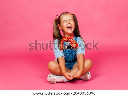 Cheerful little girl sits on the floor, rejoices and looks at the camera on a pink background.