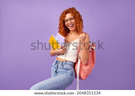 Cute redhead curly girl student teenager with a backpack holds notepads and notebooks and shows the gesture yes as a winner isolated on a purple background. Royalty-Free Stock Photo #2048351534