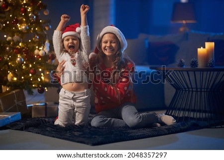 Two little caucasian sisters in funny sweaters and santa hats sitting on floor near christmas tree and laughing sincerely. Happy girls enjoying winters holidays at home. 