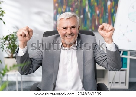 A cheerful smiling businessmen in suit with gray hair, elderly, sitting in office chair, raises hands in the air clenched in fists, the head of company won tender, signing contract, victory, happiness