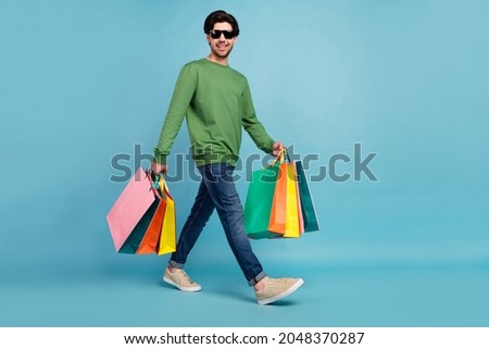 Full size photo of smiling guy wear sunglass go walk hold shopping bags shopaholic isolated on blue color background