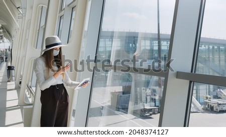 Young girl, business woman standing by the window in a modern airport holding tickets and a phone, looking at the camera. Portrait of a beautiful girl in a mask. Business trip by plane
