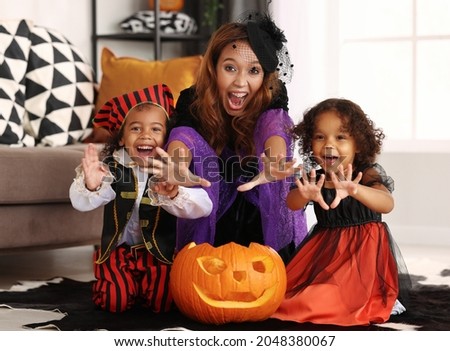 Joyful african american family mother and children in Halloween costumes making scary gesture and looking at camera while sitting on with jack-o-lantern in living room, celebrating all hallows day