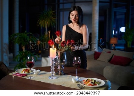 Charming lady in stylish black dress adjusting red roses in vase while preparing romantic dinner at home. Caucasian woman served table for two with glasses of wine and fresh salads. Royalty-Free Stock Photo #2048449397