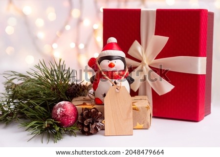A toy Santa penguin with a wooden blank form for greeting text near a red gift box with a bow, a branch of a Christmas tree on a background of blurry bokeh lights of a garland. New Year's deorations 
